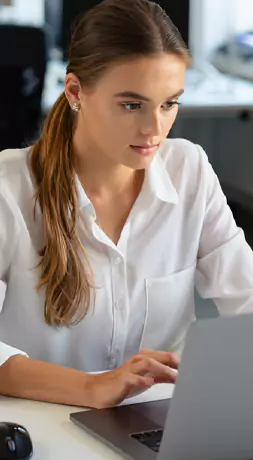 Young person in a white blouse working on a laptop at a desk.