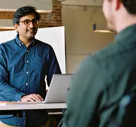 Person wearing glasses standing at a desk with a laptop, speaking to a seated colleague.